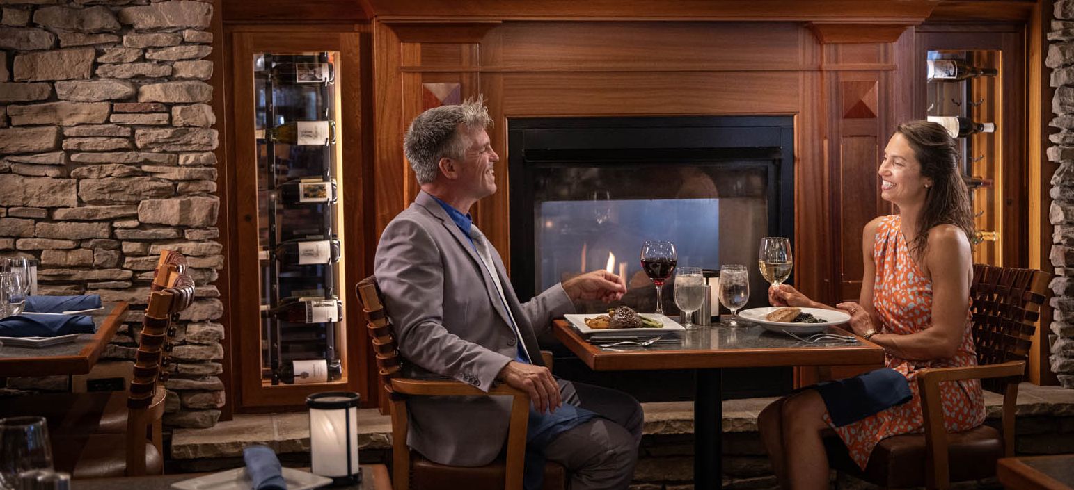 A couple in fine dress clothes enjoying a meal in front of the fireplace with mahogany trim